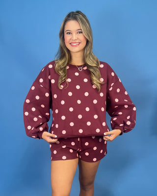 A woman modeling a matching burgundy and pink polka dot lounge set, consisting of a long-sleeve sweatshirt and shorts, against a solid blue studio background.