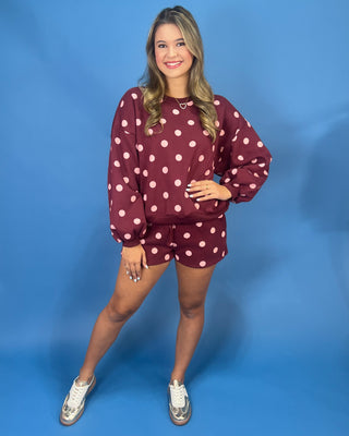 A woman modeling a matching burgundy and white polka dot sweatshirt and shorts set against a solid blue studio backdrop.