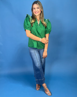 A woman modeling a vibrant green short-sleeve blouse with puffed sleeves and silver beaded trim, paired with blue jeans and metallic sandals, standing against a solid blue studio backdrop.