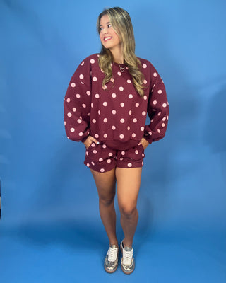 A woman modeling a matching burgundy and white polka dot sweatshirt and shorts set against a solid blue studio backdrop.