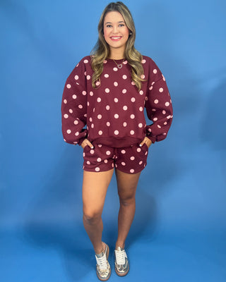 A woman modeling a matching burgundy and white polka dot sweatshirt and shorts set against a solid blue studio background.