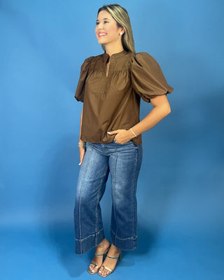 A woman modeling a brown short-sleeve blouse with puffed shoulders and a split neckline, paired with wide-leg blue jeans and metallic sandals, set against a solid blue studio backdrop.