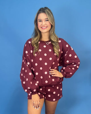 A woman modeling a matching burgundy and pink polka-dot lounge set, featuring a long-sleeve sweatshirt and shorts, against a solid blue studio background.