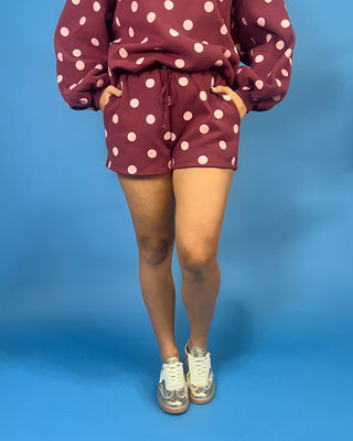 A person wearing a matching burgundy and white polka dot lounge set, consisting of a cropped sweatshirt and shorts, paired with white sneakers, against a solid blue studio background.