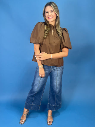 A woman modeling a brown puff-sleeve blouse with a tie-neck detail, paired with wide-leg blue jeans and metallic sandals, standing against a solid blue studio backdrop.