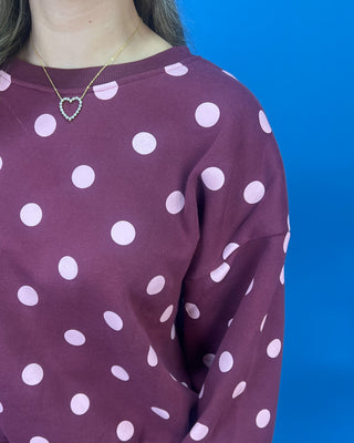 A close-up of a person wearing a long-sleeve crewneck sweatshirt in a deep plum purple with white polka dots, paired with a small heart-shaped gold necklace, set against a solid blue background.