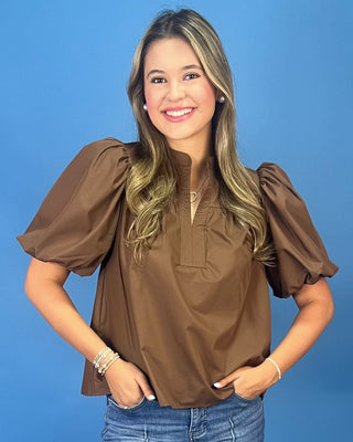 A smiling young woman with blonde-highlighted hair wearing a brown puff-sleeve blouse and blue jeans, standing against a solid blue studio background.