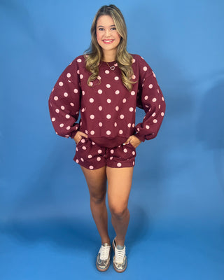 A woman modeling a matching burgundy and white polka dot lounge set, consisting of a long-sleeve sweatshirt and shorts, paired with white sneakers, against a solid blue studio backdrop.