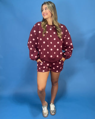 A woman modeling a matching burgundy and white polka dot sweatshirt and shorts set against a solid blue studio backdrop.