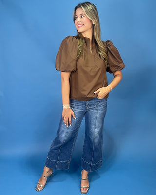 A woman modeling a brown short-sleeve blouse with puffed sleeves and a high neckline, paired with wide-leg blue jeans and metallic sandals, set against a solid blue studio background.