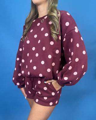 A woman wearing a matching burgundy and pink polka dot lounge set, featuring a long-sleeve sweatshirt and shorts, posed against a solid blue background.