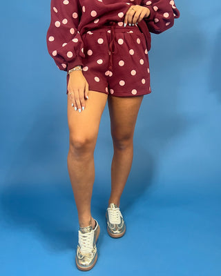 A person wearing a matching burgundy and pink polka-dot lounge set with a drawstring waist, paired with metallic silver sneakers, against a solid blue studio background.