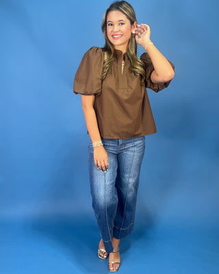 A woman modeling a brown puff-sleeve blouse with a split neckline and front button detail, paired with blue jeans and metallic sandals, standing against a solid blue studio backdrop.