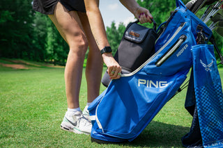 Person opening a blue golf bag with 'PING' branding on a grassy field.