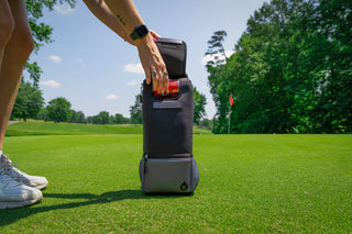 Person using a golf trolley on a golf course with trees and a clear sky in the background.