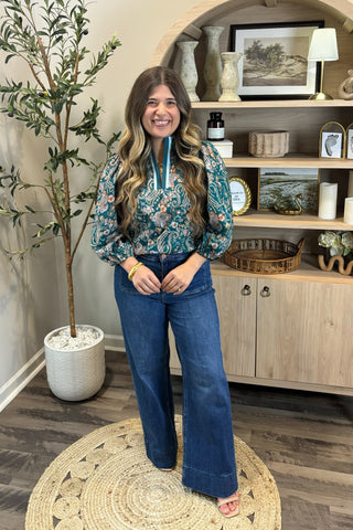 Woman in a floral blouse and jeans standing in a room with a plant and shelves.
