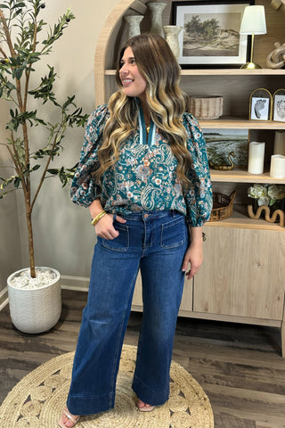 Woman wearing a floral blouse and blue jeans standing in a room with decor elements.