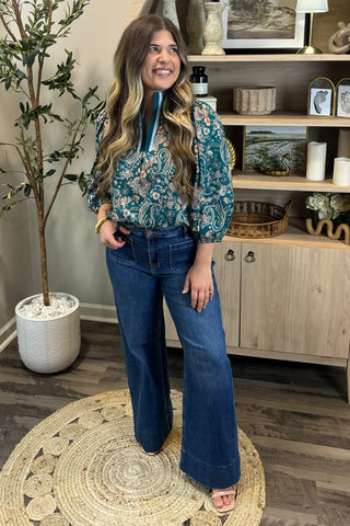 Woman in a teal patterned blouse and blue jeans standing in a room with a wooden cabinet and plant.