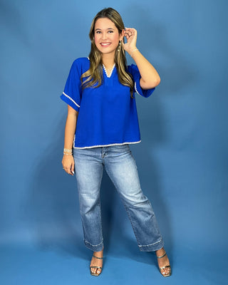 A woman modeling a bright royal blue short-sleeve top with white trim, paired with light-wash jeans and metallic sandals, standing against a solid blue studio backdrop.