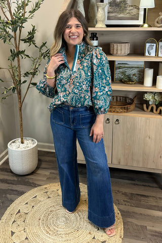 Woman in a floral blouse and blue jeans standing in a room with a plant and shelves.