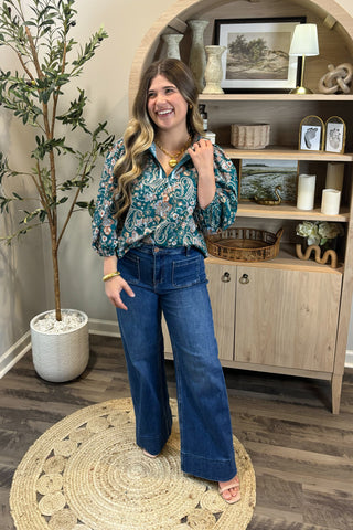 Woman in a floral blouse and blue jeans standing in a room with a wooden cabinet and decorative items.