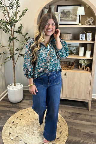 Woman in a floral blouse and blue jeans standing in a room with wooden flooring and a decorative shelf.