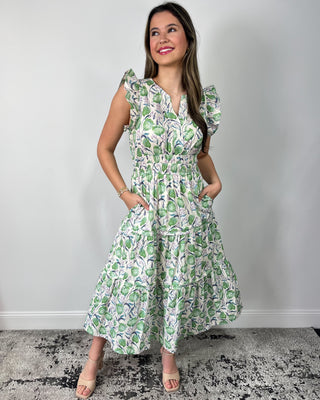 Smiling woman posing in front of a neutral backdrop wearing a breezy, tiered midi dress with a soft green botanical print and flutter sleeves, featuring a flattering cinched waist and flowy skirt for effortless movement, styled with nude heeled sandals and simple gold jewelry for a polished, garden-party ready look.