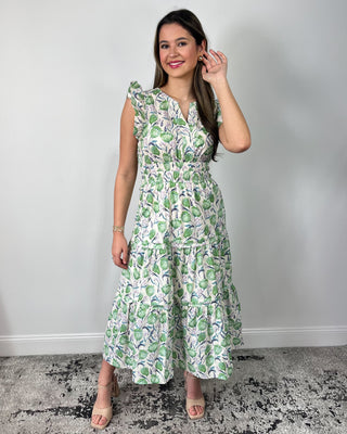 Smiling woman posing in front of a neutral backdrop wearing a breezy, tiered midi dress with a soft green botanical print and flutter sleeves, featuring a flattering cinched waist and flowy skirt for effortless movement, styled with nude heeled sandals and simple gold jewelry for a polished, garden-party ready look.