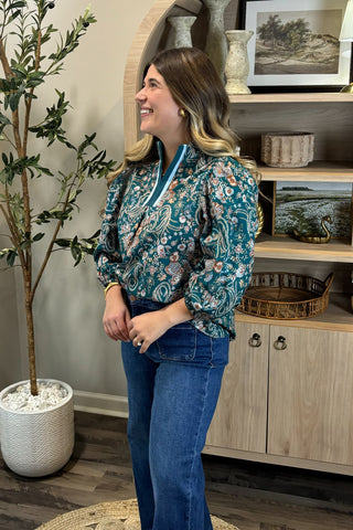 Woman in a floral blouse and jeans standing in a room with a plant and wooden cabinet.