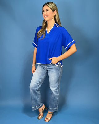 A woman modeling a bright royal blue short-sleeve blouse with white trim, paired with light-wash jeans and metallic sandals, standing against a solid blue studio backdrop.