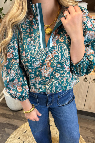 Person wearing a floral blouse with a gold necklace and blue jeans indoors.