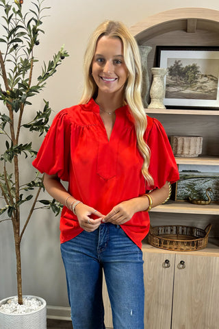 Woman wearing a red blouse and blue jeans standing in a room with a plant and shelves.