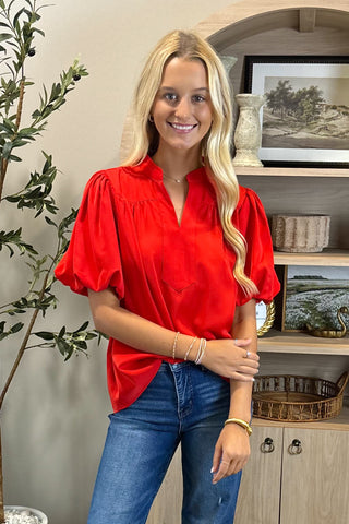 Woman wearing a red blouse and blue jeans standing in a room with a shelf and plant.