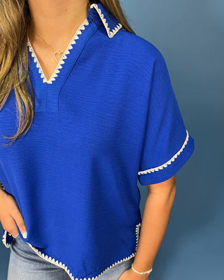 A woman wearing a bright royal blue short-sleeve top with white scalloped trim along the V-neckline and sleeve cuffs, paired with light blue jeans against a solid blue-gray background.