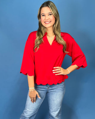 A woman modeling a bright red V-neck blouse with three-quarter bell sleeves and a scalloped hem, paired with light-wash blue jeans, against a solid blue studio background.