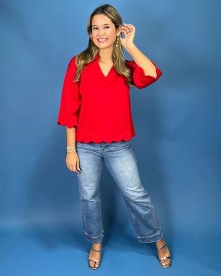 A woman modeling a bright red blouse with three-quarter ruffled sleeves and a scalloped hem, paired with light-wash blue jeans and metallic sandals, standing against a solid blue studio backdrop.