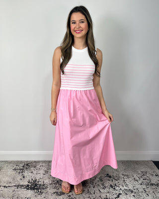 Smiling woman posing in front of a neutral backdrop wearing a sweet and feminine outfit featuring a sleeveless white ribbed tank with soft pink horizontal stripes paired with a flowy high-waisted pink maxi skirt, styled with clear slide sandals and simple gold jewelry for an effortless, spring-ready look.
