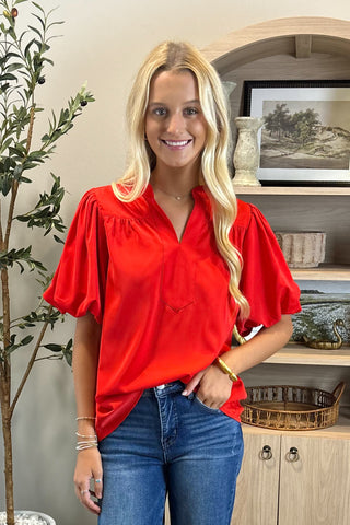 Woman wearing a red blouse and blue jeans standing in a room with a plant and shelves.