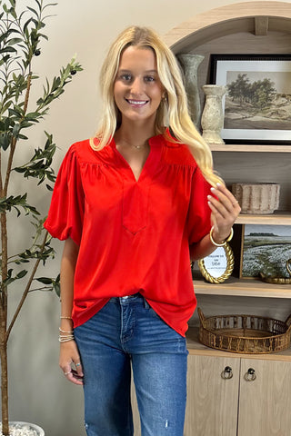 Woman wearing a red blouse and blue jeans standing in a room with decor elements.