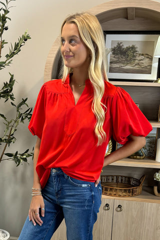 Woman wearing a red blouse and blue jeans standing in a room with a plant and cabinet.