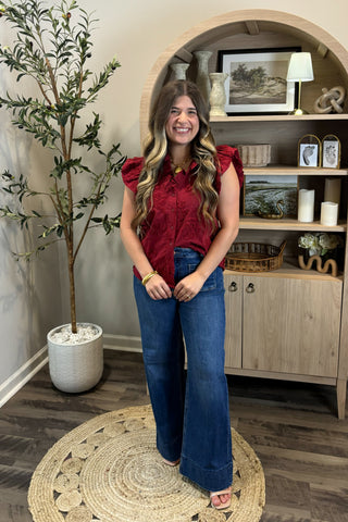 Woman in a red top and blue jeans standing in a room with a plant and wooden cabinet.