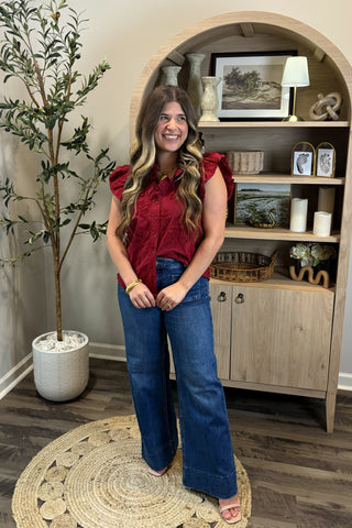 Woman in a red top and blue jeans standing in a room with a wooden cabinet and decorative items.