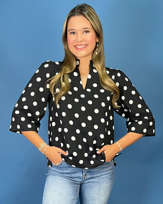 A woman modeling a black blouse with white polka dots, paired with light blue jeans, standing against a solid blue studio background.