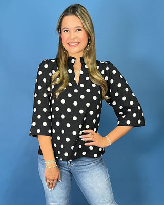 A woman modeling a black three-quarter sleeve blouse with white polka dots and a split neckline, paired with light blue jeans, set against a solid blue studio background.
