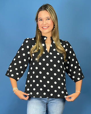 A woman modeling a black three-quarter sleeve blouse with white polka dots, paired with light blue jeans, against a solid blue studio background.