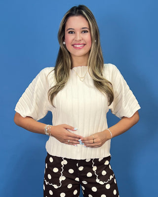 A smiling woman with long blonde-highlighted hair stands against a solid blue studio background, wearing a cream ribbed short-sleeve top and dark brown pants with large white polka dots.