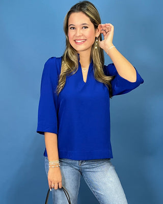 A woman modeling a bright royal blue short-sleeve blouse with a split neckline, paired with light-wash blue jeans, standing against a solid blue studio backdrop.
