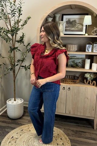 Woman in a red blouse and blue jeans standing in a room with a plant and wooden cabinet.