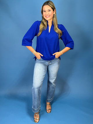A woman modeling a royal blue three-quarter sleeve blouse with a split V-neckline, paired with light-wash blue jeans and metallic sandals, standing against a solid blue studio backdrop.