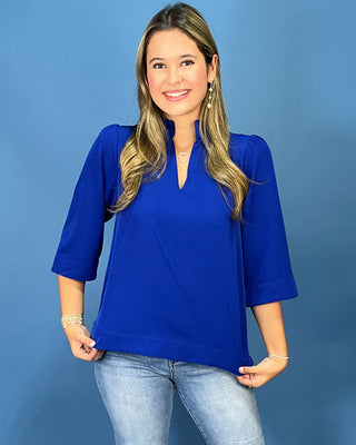 A woman modeling a bright royal blue three-quarter sleeve top with a split neckline, paired with light-wash blue jeans, set against a solid blue studio background.